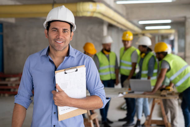 Portrait of cheerful architect at a construction site holding a clipboard smiling at camera Portrait of cheerful architect at a construction site holding a clipboard smiling at camera - Incidental people working at background civil engineer stock pictures, royalty-free photos & images