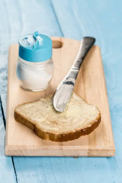 Flat lay butter spread on bread with knife and saltshaker served on a wooden board.