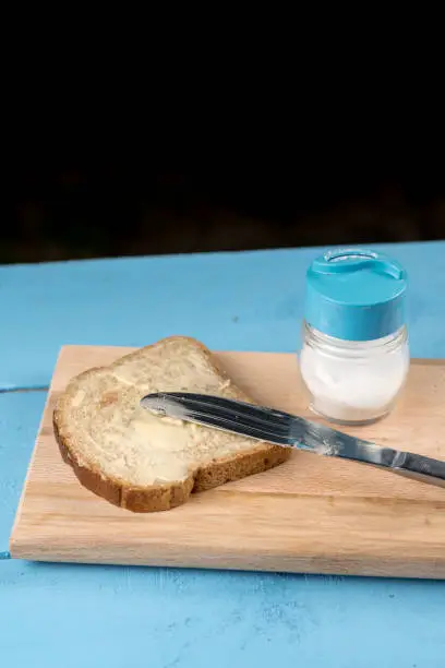 Flat lay butter spread on bread with knife and saltshaker served on a wooden board.