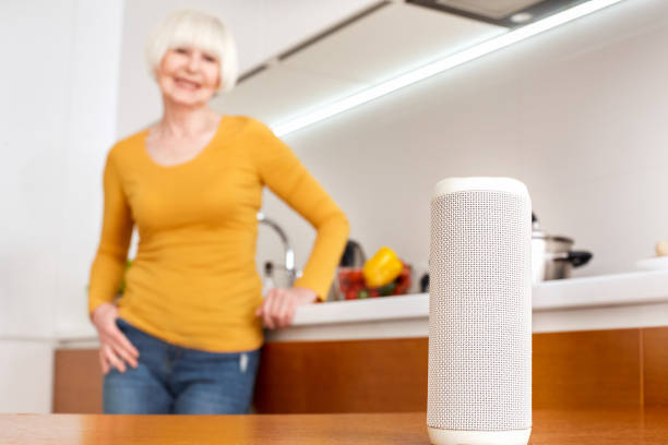 istock Smart AI white color audio speaker concept. Mini voice assistant helper at home in foreground of old beauty woman at kitchen. Portrait of senior use modern technology innovation siri device. Care 1219462058