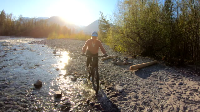 Mature woman bikes through river shallows, mountains