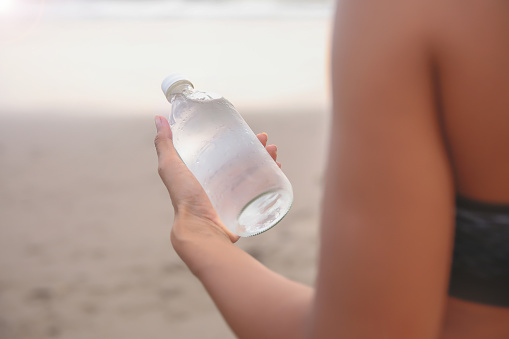 Close up shot of Asian woman holding water bottle on her hand to drink while walking on empty beach, during Covid 19 pandemic
