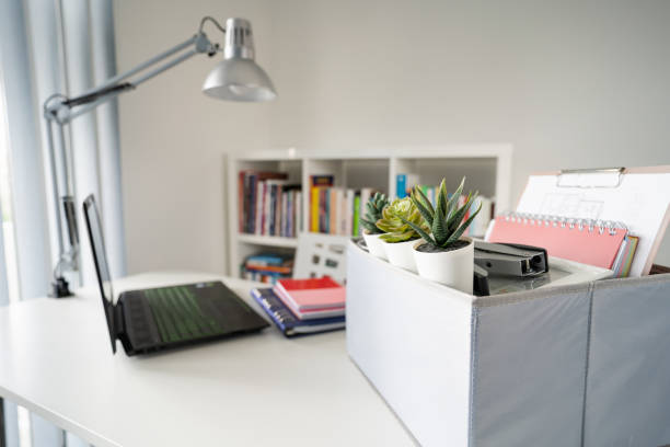 istock Box with personal items stuff packed on the white table in the office or home in day by the laptop ready to move after being fired or got the job at work place side view 1219477419