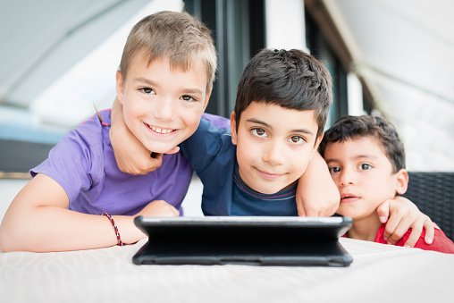 Small group of young boys playing together with a digital tablet. Hugging each other. Looking towards the camera. Young schoolboys with electronic gadgets lifestyle portrait.