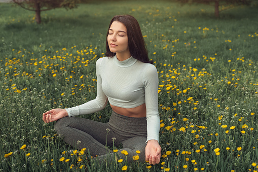 Woman meditating in lotus pose on green dandelion meadow against forest. Practicing of yoga at summer. Training and meditation outdoor. Healthy lifestyle. Woman enjoying sunny springtime day in meadow.