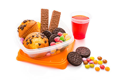 High angle view of a school lunch box filled with unhealthy food for kids isolated on white background. High sugar and fat  levels like muffins, chocolate bars, cookies, jellybeans and a cola glass are included in the composition. High resolution 42Mp studio digital capture taken with SONY A7rII and Zeiss Batis 40mm F2.0 CF lens