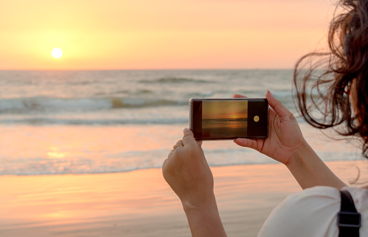 Hermosa foto de vista lateral de joven turista explorando el mundo durante las vacaciones y está tomando una foto de puesta de sol / amanecer en su teléfono inteligente. La foto a hacer clic es visible en el visor del teléfono. photo