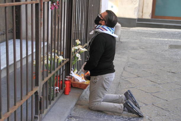 Catholic faith at the time of churches closed for health emergency Pagani,Sa,Italy - April 17,2020 :A faithful Catholic on his knees in front of the closed gate of the Shrine of Our Lady of the Chickens . A prayer in silence and gifts brought to Our Lady. Churches closed cause of emergency from covid-19 buddhist prayer wheel stock pictures, royalty-free photos & images