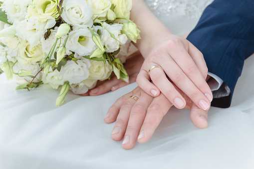 Bride and groom hold hands with golden wedding rings next to wedding bouquet. Shallow depth of field.