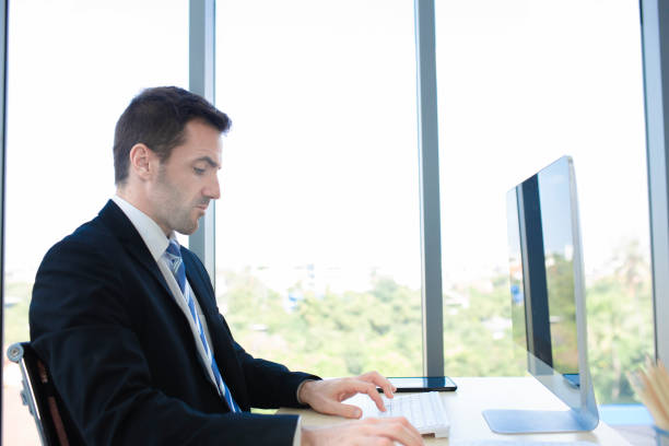 istock Side view handsome young businessman in eyewear working with computer remotely, sitting at wooden table in kitchen. Pleasant happy man communicating in social network, searching information online.