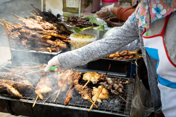 Photo of Thai style Isaan street food vendor cart,grilled chicken,catfish and grilled chicken liver on a charcoal stove with smoke at papaya salad(Som Tam Thai)famous local Thai street food hawker stall