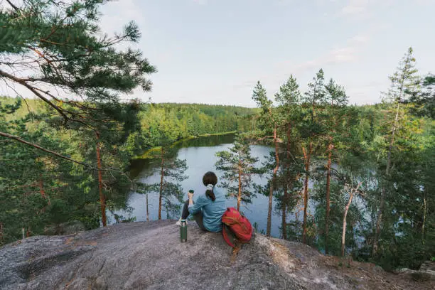 Young Caucasian woman sitting, looking at lake in Finland and drinking tea