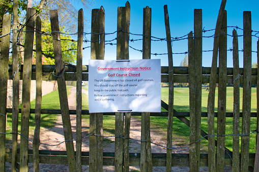 In dappled light on a sunny day, a paper sign in plastic is attached to the locked gates at a suburban golf course in Merseyside in the UK. The sign warns that the course is closed due to the UK Government lockdown to control the global coronavirus pandemic.