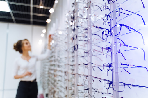 Eyeglasses shop. Stand with glasses in the store of optics. Woman on blurred background chooses glasses.