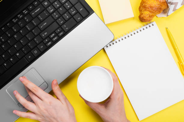 istock Working morning. Girl works for laptop. Breakfast at work - cappuccino, croissant, notebook on yellow background. Top view 1219528621