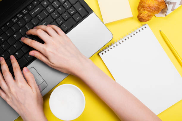 istock Working morning. Girl works for laptop. Breakfast at work - cappuccino, croissant, notebook on yellow background. Top view 1219528642