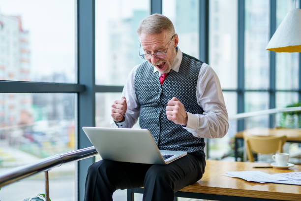 glücklicher geschäftsmann im anzug blick auf laptop aufgeregt von guten nachrichten online, glücklich enise erfolgreichen gewinner mann sitzt am büro schreibtisch und hebt hand in ja geste feiern geschäftserfolg gewinnen ergebnis - compiling stock-fotos und bilder