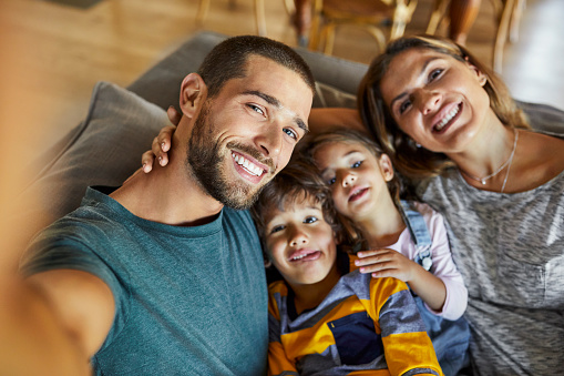 High angle view of smiling man taking selfie with family. Portrait of parents and children sitting together on sofa in living room. They are having fun during quarantine at home.