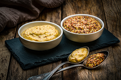 Spices: close up view of two bowls filled with whole grain and Dijon mustard shot on dark wooden table. Spoons with mustard are in front of the bowls. Predominant colors are gold and brown. High resolution 42Mp studio digital capture taken with Sony A7rII and Sony FE 90mm f2.8 macro G OSS lens