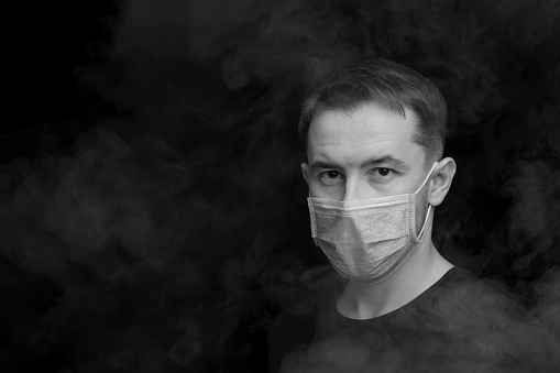 Dark black and white image. Caucasian adult man in protective mask surrounded by smoke. Horizontal orientation, copy space. Air pollution concept