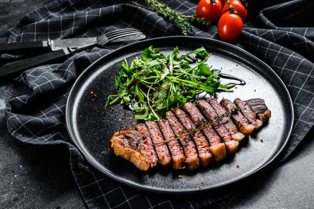 istock Cooked grilled strip loin steak, marbled beef meat with arugula. Black background. Top view 1219555898