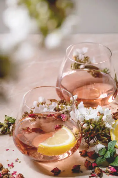 Cocktail glasses of pink rose champagne, cider or lemonade with dry rose buds, lemon and mint. Blossom cherry branches above. Pink marble background.