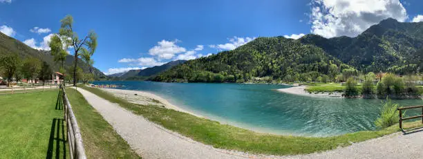 Panorama of coast of lake Molveno, the Italian Alps, quiet water, no people, reflection of clouds in water, a pier, green trees and mountains on a background