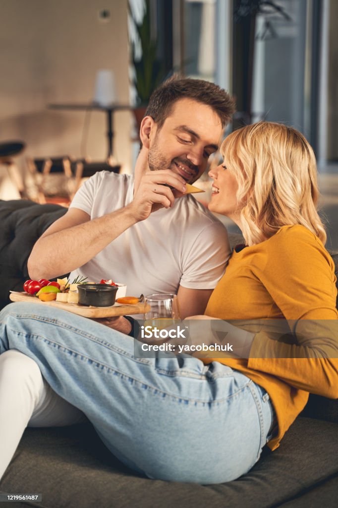 Happy married people eating together on the sofa Loving young husband feeding his hungry wife with a slice of Parmesan cheese at breakfast Eating Stock Photo