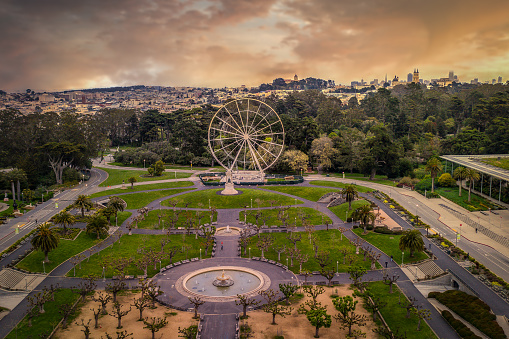 An aerial view of the new ferris wheel in Golden Gate park at sunrise with a golden sky. The San Francisco skyline is seen in the distance.