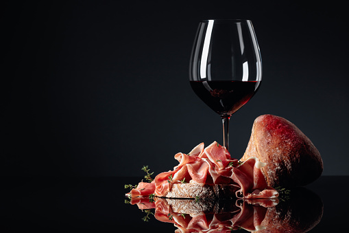 Prosciutto with ciabatta, red wine and thyme on a black reflective background. Copy space.