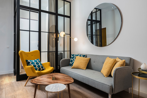 Sala de estar colorida y acogedora con sillón y sofá de diseño, junto con un espejo decorativo redondo y una pared de cristal. photo