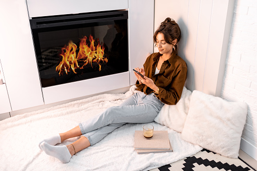 Young woman relaxing with a smart phone, sitting near the fireplace at the modern living room at home
