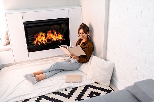 Young woman reading book, relaxing near the fireplace at the modern living room at home