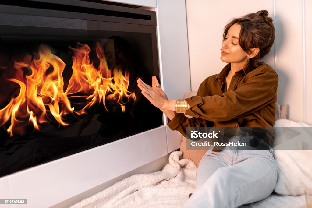 Woman relaxing near the fireplace at home Young woman relaxing near the fireplace, warming hands at the modern living room at home Adult Stock Photo