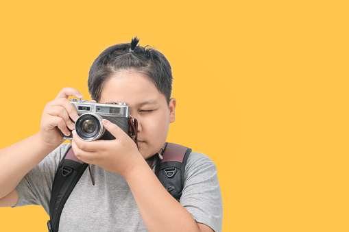 Obese boy traveler taking a picture with a vintage camera looking through the viewfinder isolated on yellow background with copy space and clipping path