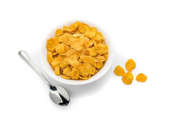 Top view of a ceramic bowl filled with crunchy corn flakes isolated on white background. A green plastic spoon is beside the bowl. Predominant colors are yellow and white. High resolution 42Mp studio digital capture taken with SONY A7rII and Zeiss Batis 40mm F2.0 CF lens
