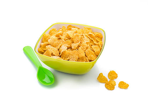 Green bowl filled with crunchy corn flakes isolated on white background. A green plastic spoon is beside the bowl. Predominant colors are yellow, green and white. High resolution 42Mp studio digital capture taken with SONY A7rII and Zeiss Batis 40mm F2.0 CF lens