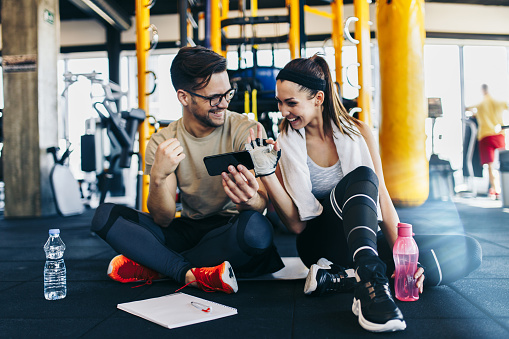 Male fitness gym instructor resting and sitting with young attractive woman on floor while making workout plan on his smart phone.