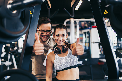 Young attractive couple working out smiling and having fun in modern fitness gym. They showing thumb up sign.