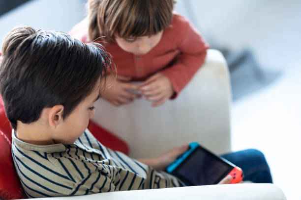 mały chłopiec grający w gry wideo siedząc na kanapie, a jego brat patrzy mu w domu. - playing video game little boys playful zdjęcia i obrazy z banku zdjęć