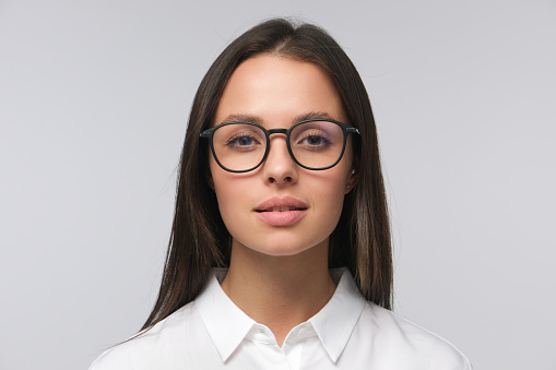 Portrait of young business woman in white collar shirt and glasses, looking straight at camera attentively with neutral smile, isolated on gray background