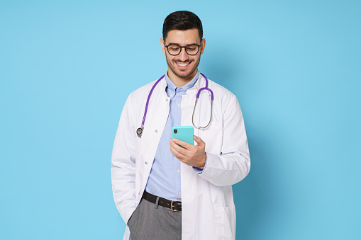 Cheerful young doctor in white coat smiling while looking at screen of his phone, using medical app, standing isolated on blue background