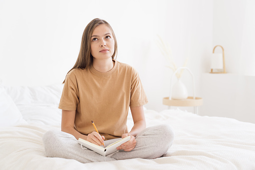 Young woman spending free time in bedroom, sitting on bed cover, thinking of plans for week and writing them in notebook that is on her knees