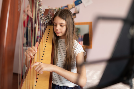 Girl playing harp at home. She is practicing playing her instrument in children room.\nPhoto taken during quarantine because of corona virus outbreak.