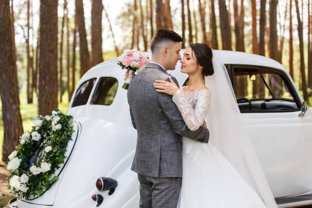 elegante brautpaare, bräutigam im grauen anzug und braut im brautkleid mit langen ärmeln und langem schleier, der sich in der nähe von weißen just married auto - veil bride lace married stock-fotos und bilder