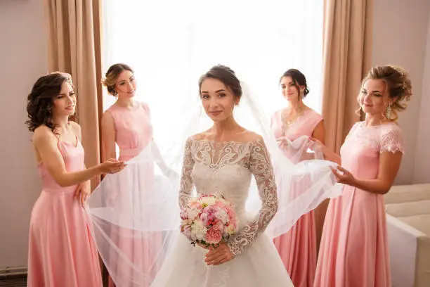 Bride in white wedding dress with long sleeves and bridesmaids in pink dress posing near window before wedding ceremony