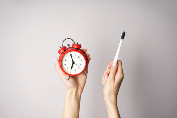 Red alarm clock and a toothbrush in the hands of a young woman on a gray background. Top view. Morning concept stock photo