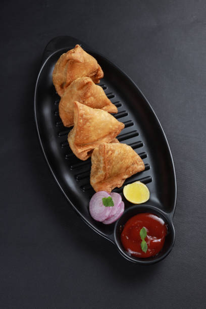 Samosa Indian samosas - fried/baked pastry with savoury filling, popular Indian snacks, served in black dish with sauce fresh onion and lemon cilantro on rustic background. masala stock pictures, royalty-free photos & images