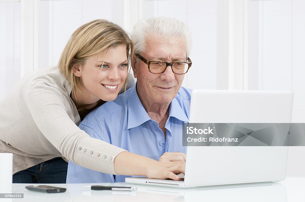 Young woman helping older man on laptop Happy smiling young girl teaching and showing new computer technology to her grandfather. Senior Men Stock Photo