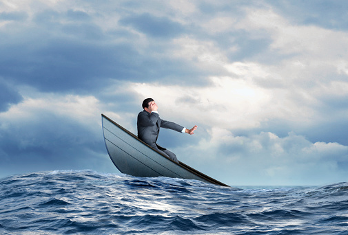 A fearful businessman places his hand over his eyes as the boat he is on is about to sink into the ocean.